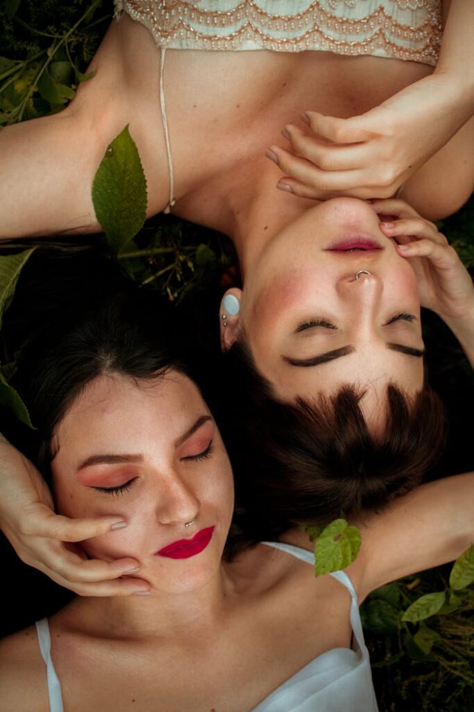 Top view of serene young females with bright makeup lying on grassy ground with eyes closed head to head and touching each others face gently